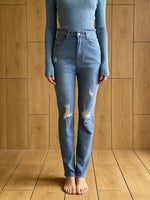 Person wearing a blue ribbed long-sleeve top and distressed blue jeans standing against a wooden paneled wall.