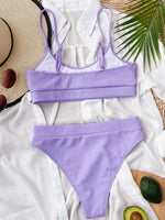 Lavender bikini set on a white fabric background with a straw hat and avocado.