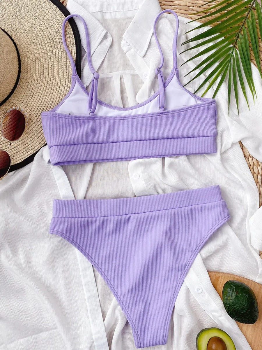 Lavender bikini set on a white fabric background with a straw hat and avocado.