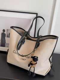 Beige tote bag with black handles and a small dog plushie on a dark surface.