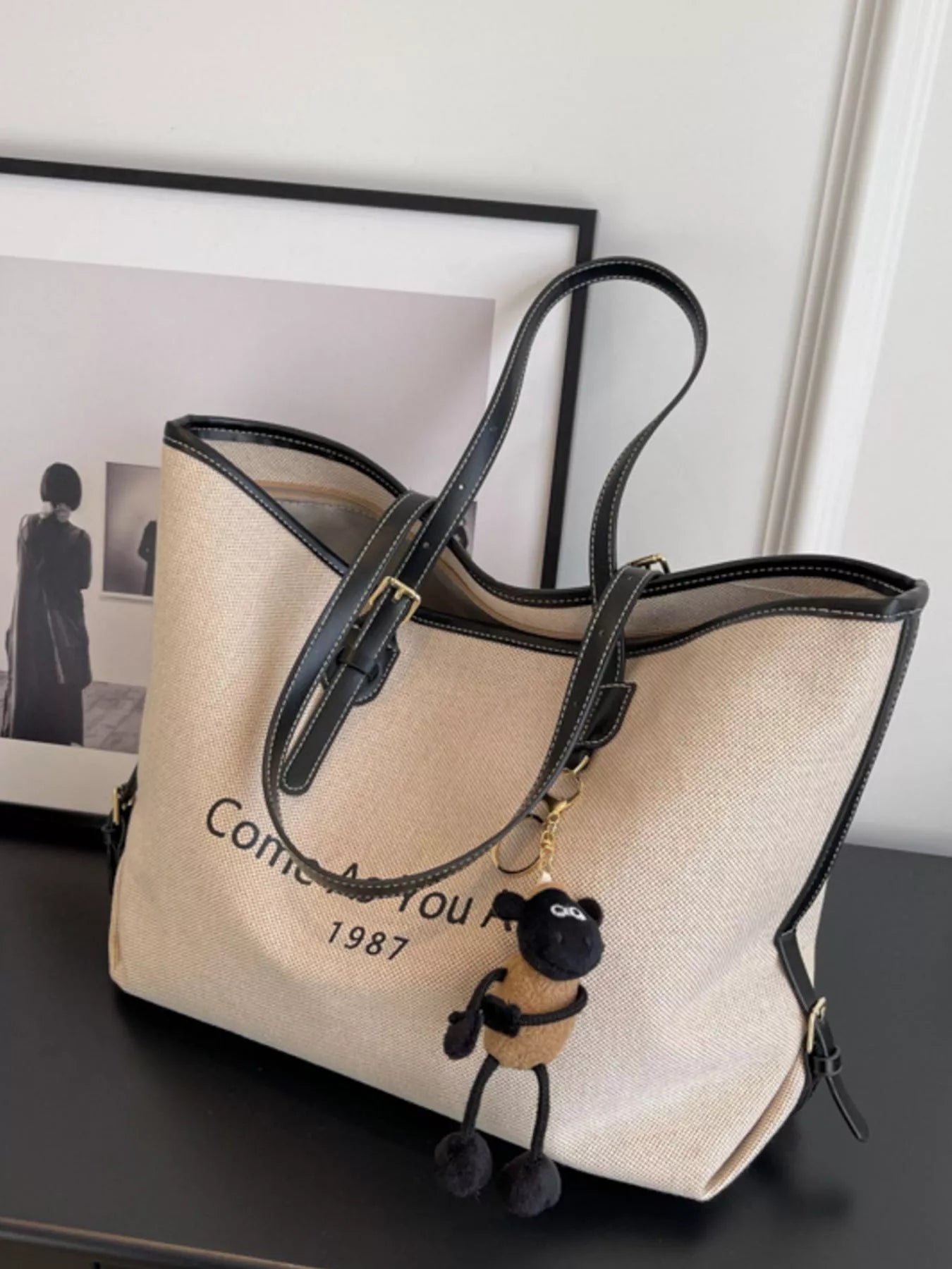 Beige tote bag with black handles and a small dog plushie on a dark surface.