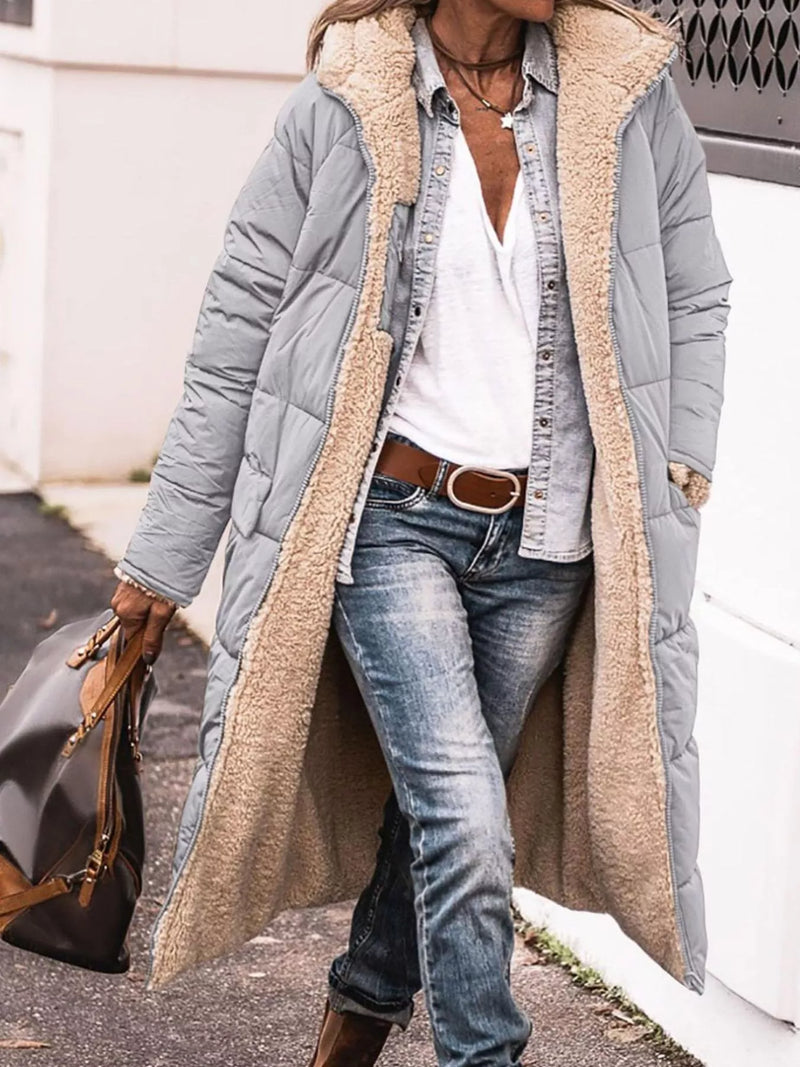 A woman wearing a long light gray puffer coat with a beige sherpa lining, styled with a white shirt, denim jacket, blue jeans, and brown boots, while carrying a dark brown handbag.