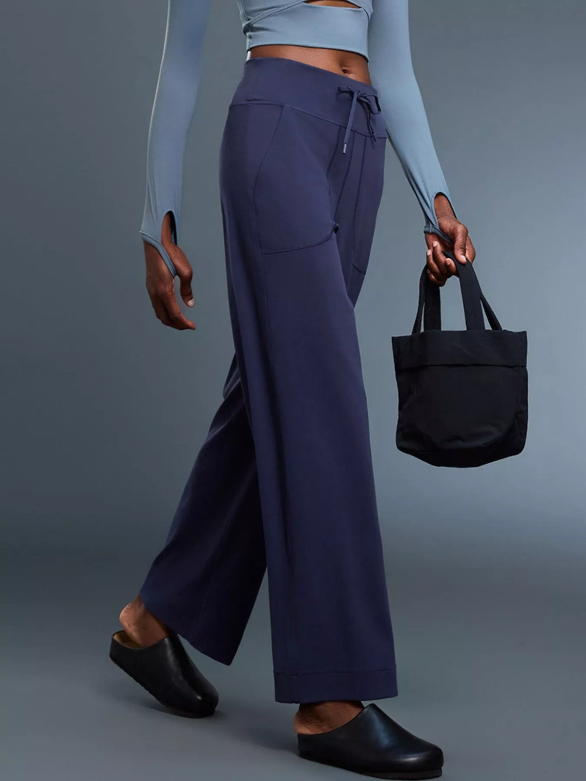 Person wearing navy pants and a light blue top, holding a black tote bag on a gray background