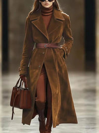 Woman wearing a long brown coat with a belt, holding a matching handbag, against a blurred background.