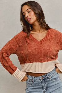Woman wearing a rust-colored cable knit sweater with a beige trim and blue jeans against a gray background