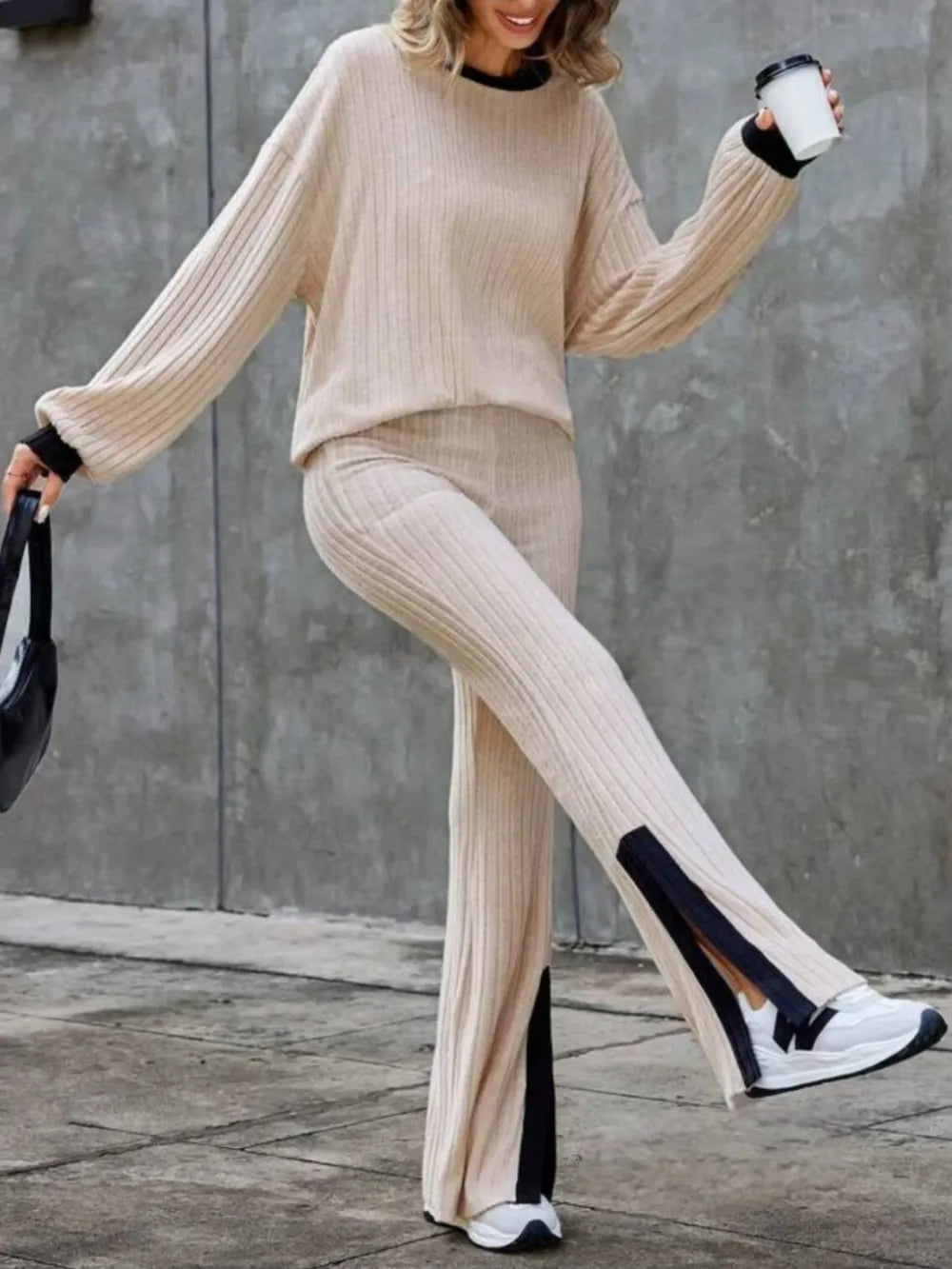 Person wearing a beige ribbed sweater and pants with black accents, holding a coffee cup.