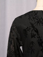 Close-up of the back of a black sheer embroidered blouse or dress with floral and bird motifs, shown on a mannequin against a light gray backdrop.