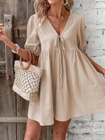 Woman wearing a beige dress holding a woven bag against a rustic wooden door and white brick wall.