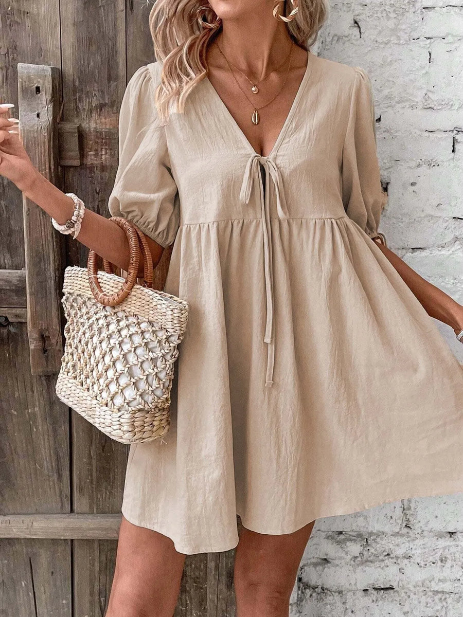 Woman wearing a beige dress holding a woven bag against a rustic wooden door and white brick wall.