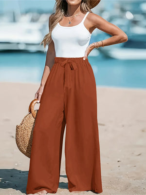 Woman in white top and brown wide-leg pants standing on a beach with boats in the background.