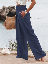 Person wearing navy wide-leg pants with a beige bag by a body of water.