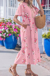Woman in a pink floral dress holding a woven bag, standing in front of colorful flowers and a building.