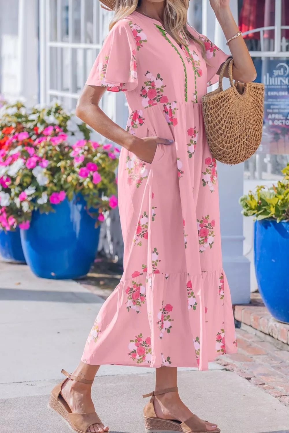 Woman in a pink floral dress holding a woven bag, standing in front of colorful flowers and a building.