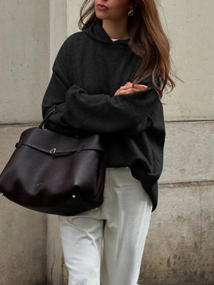 Person wearing a black oversized sweater and holding a black leather bag against a concrete wall.
