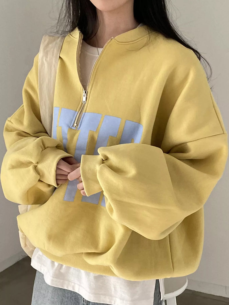 A person wearing an oversized yellow half-zip sweatshirt with a large light blue graphic print on the front, layered over a white top and paired with light blue jeans.