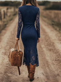 Woman in a blue lace dress and brown boots walking on a dirt road with a leather bag.