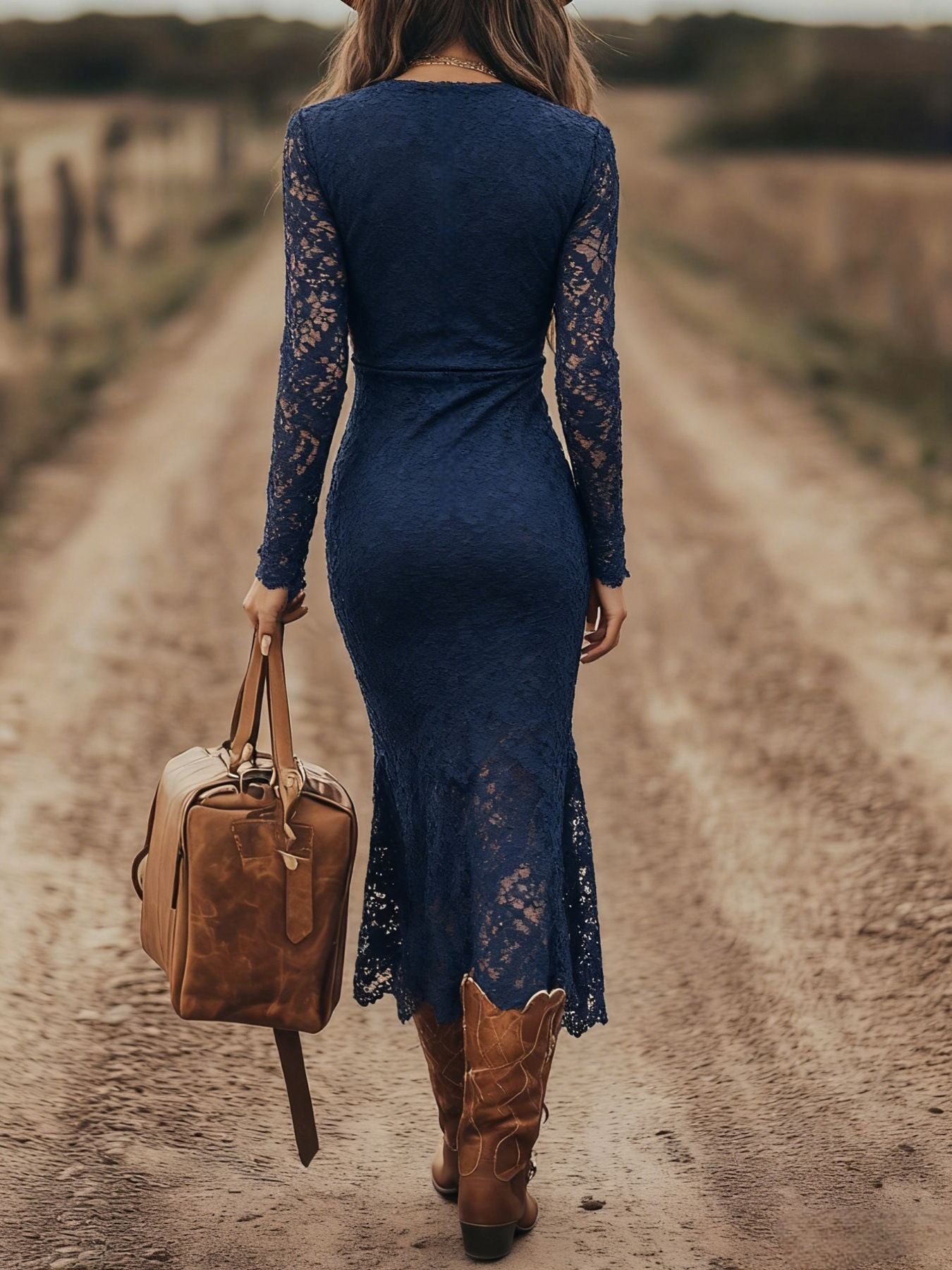 Woman in a blue lace dress and brown boots walking on a dirt road with a leather bag.