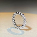 A silver-toned eternity ring set with clear round-cut gemstones, photographed on a warm beige surface with a soft shadow and a blurred gray background.