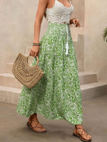 Woman wearing a green patterned dress with a white top, holding a woven bag on a neutral background.