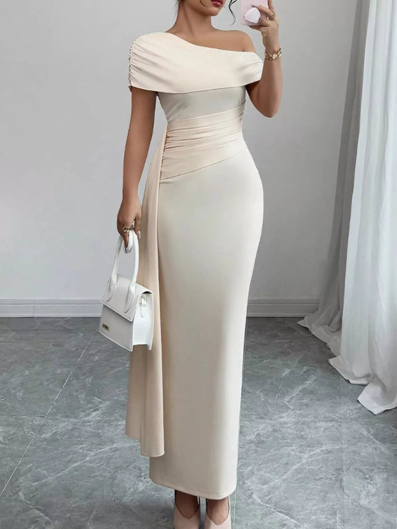 Woman wearing a beige off-shoulder dress holding a white handbag indoors.
