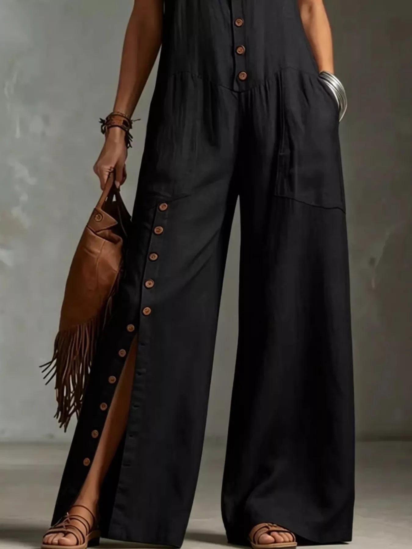 Person wearing a black jumpsuit with button details, holding a brown leather bag against a neutral background.