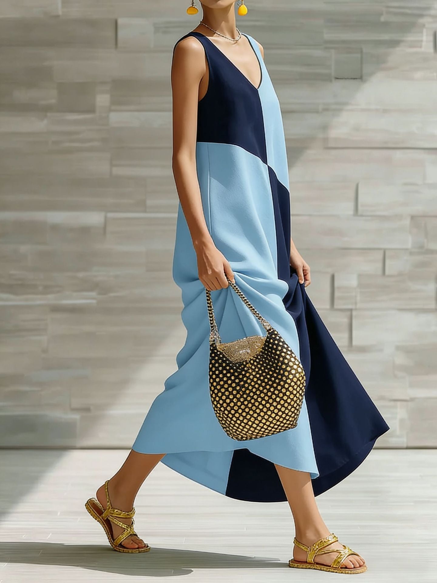 Woman in a blue and navy dress holding a patterned handbag against a stone wall.