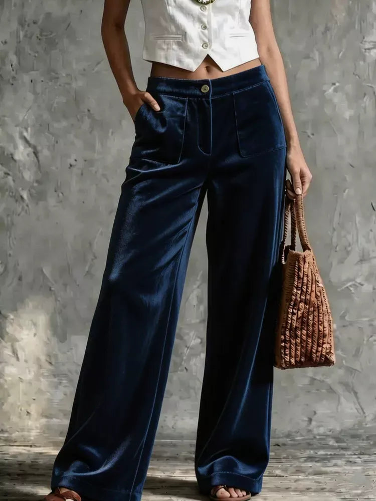 A person wearing high-waisted, wide-leg navy blue velvet pants, styled with a white cropped top and holding a brown woven handbag against a textured gray wall.