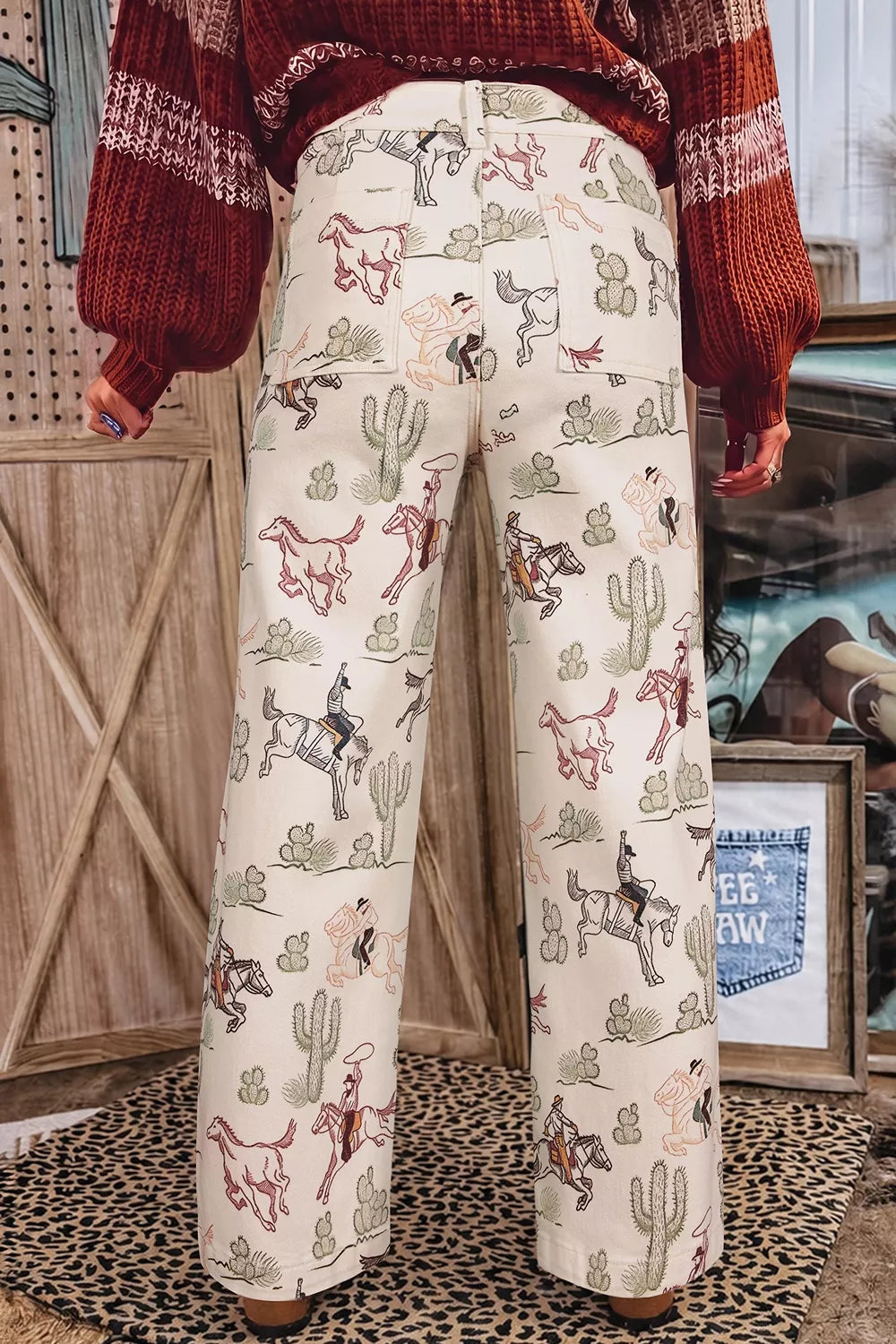 A person shown from the back wearing wide-leg cream pants with a western print of cacti, horses, and riders, paired with a rust red knit sweater in a rustic indoor setting.