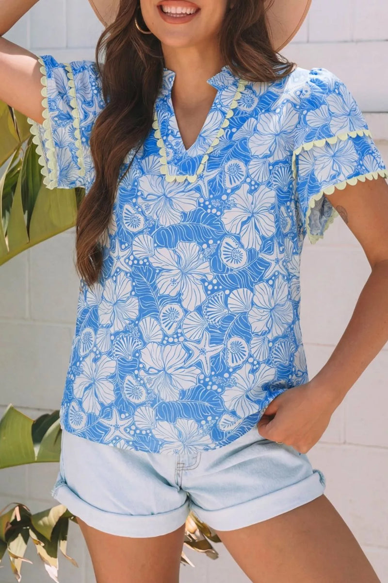 Woman wearing a blue floral blouse with white accents and light blue shorts, standing against a white tiled wall.