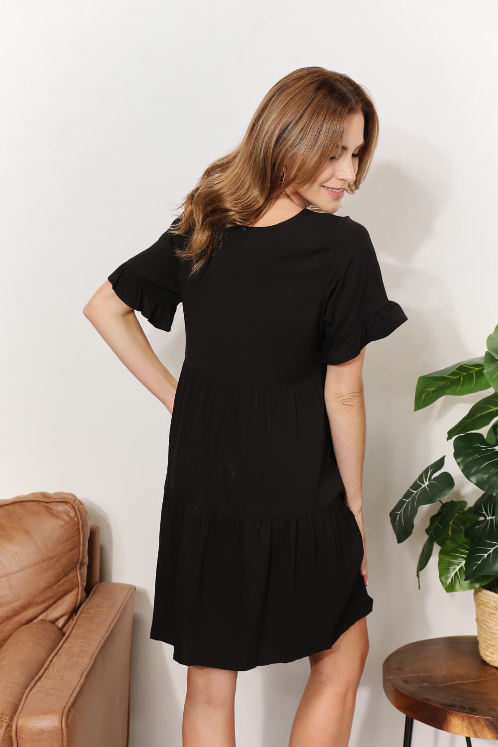 Woman wearing a black dress standing in a room with a plant and leather chair.