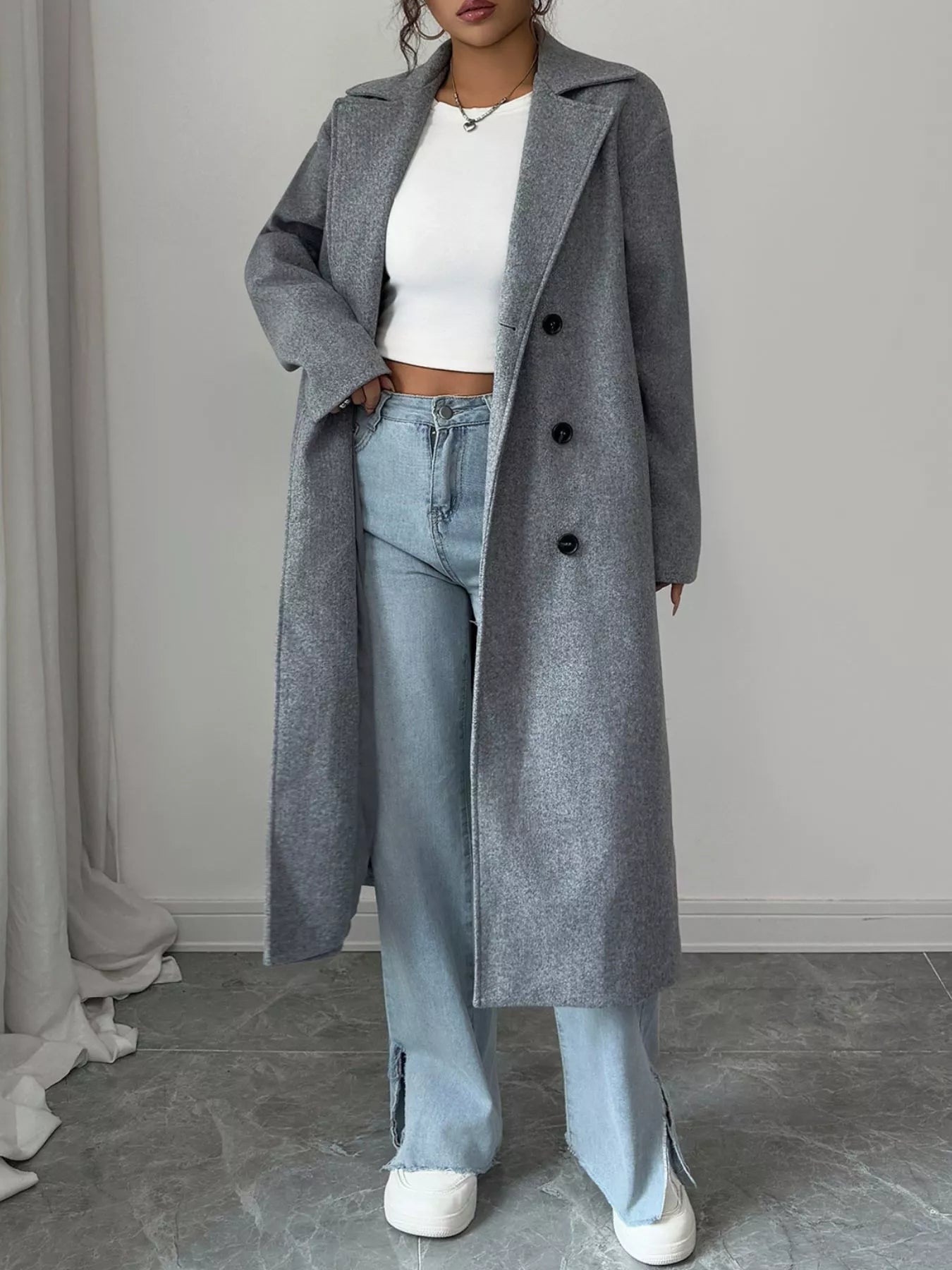 Person wearing a long gray coat, white top, and light blue jeans against a plain background
