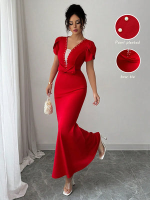 Woman wearing a red dress with pearl pleated details and bow tie, standing in a room with white curtains.