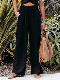 Person wearing black wide-leg pants and a black top, holding a beige handbag in front of plants.