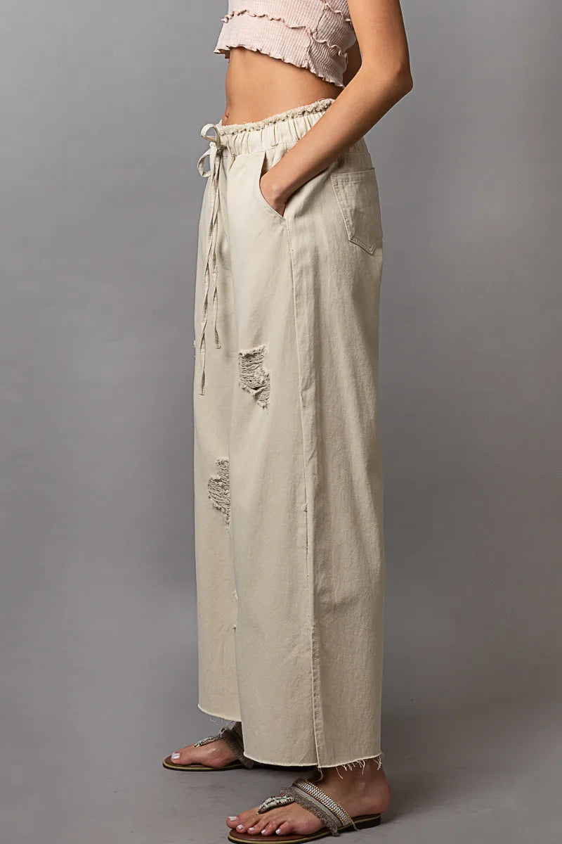 Beige wide-leg pants with distressed details worn by a person on a gray background