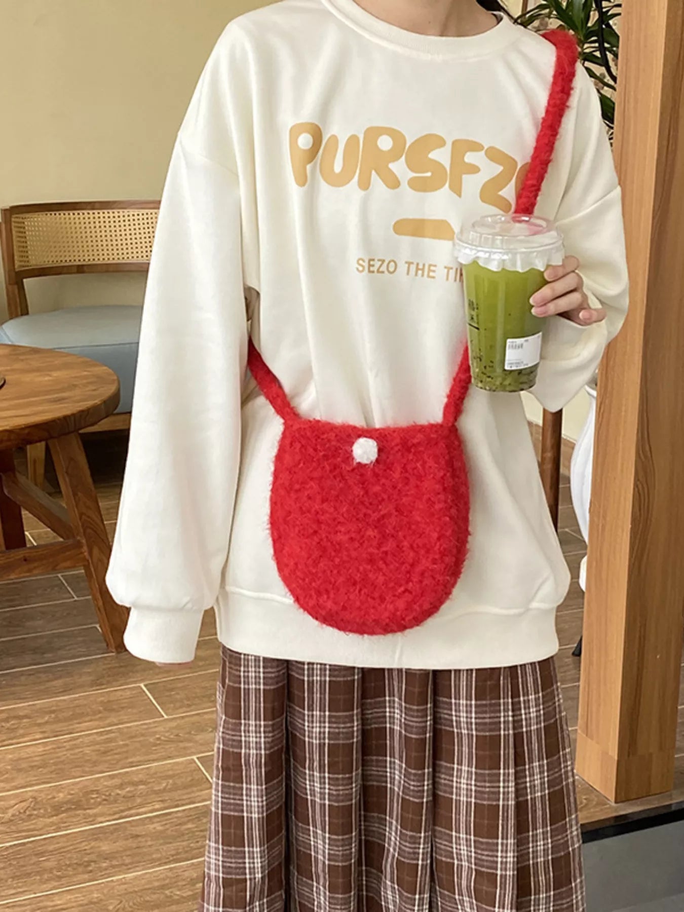 Person wearing a white sweatshirt with 'PURSFZ' text, holding a red bag and a green drink.