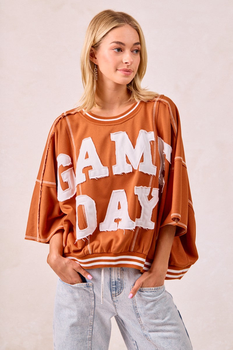 Woman wearing a 'GAME DAY' brown oversized shirt with light blue jeans on a beige background
