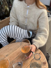 Person sitting at a table with a glass of orange liquid, wearing a beige sweater and striped pants.