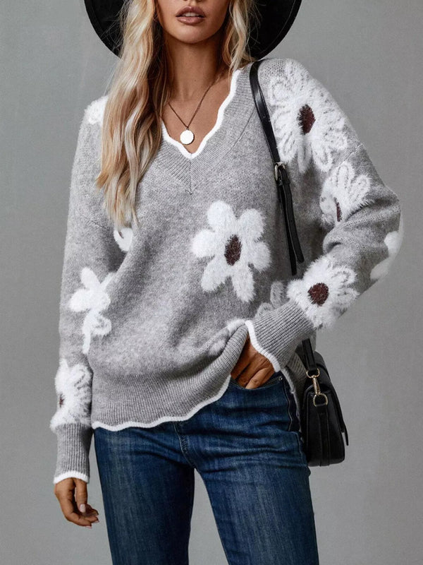 A woman wearing a gray knit sweater with large white daisy-like floral patches and white scalloped trim, paired with blue jeans and a black hat, against a plain gray studio background.