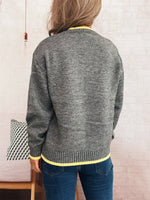 Person wearing a gray sweater with yellow trim, standing against a light-colored wall.