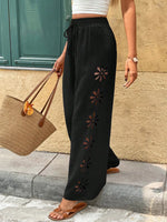 Person wearing black pants with floral cutouts, holding a brown bag against a beige wall.