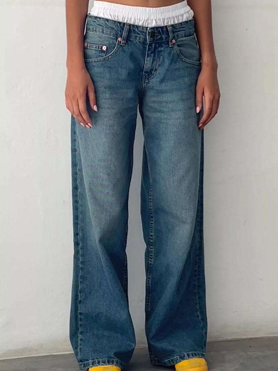 A person wearing medium-wash blue wide-leg jeans, shown from the waist down against a plain off-white wall.