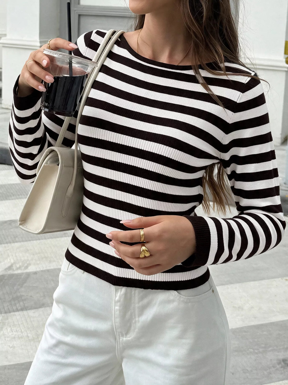 A woman wearing a black and white horizontally striped long-sleeve knit top, paired with white pants and carrying a light beige shoulder bag.