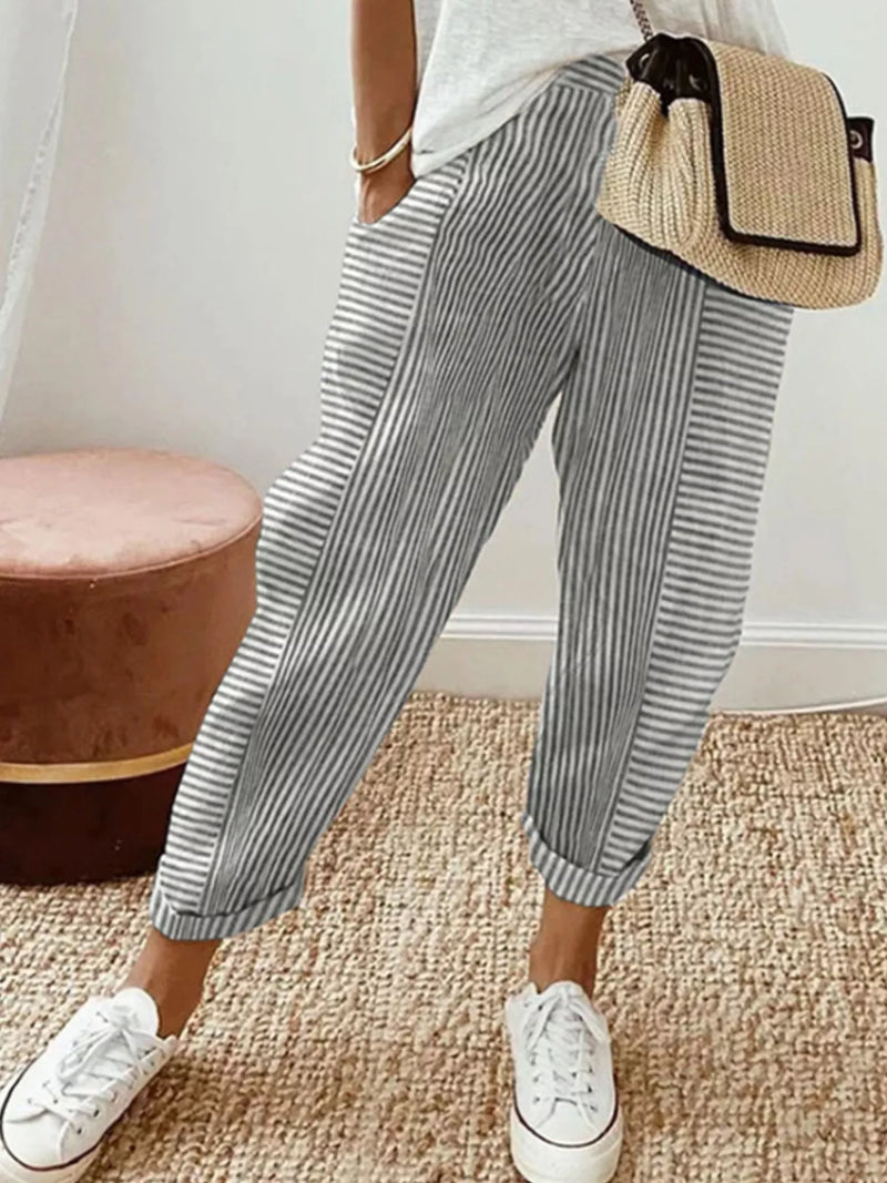 A person wearing loose, cuffed striped pants in gray and white, paired with white sneakers and a woven straw crossbody bag, standing on a textured beige rug.