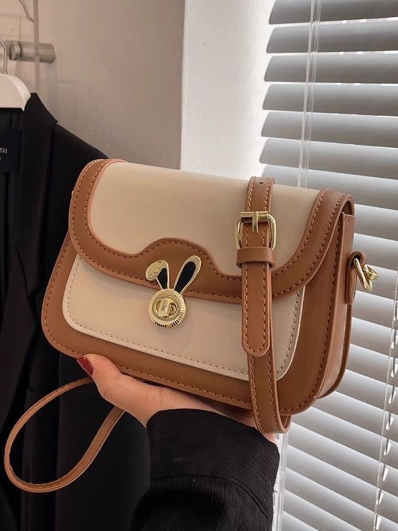 Beige and brown handbag with a gold buckle held by a person in front of a window with blinds.