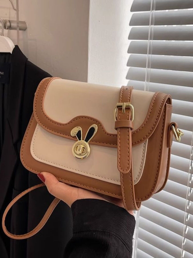 Beige and brown handbag with a gold buckle held by a person in front of a window with blinds.