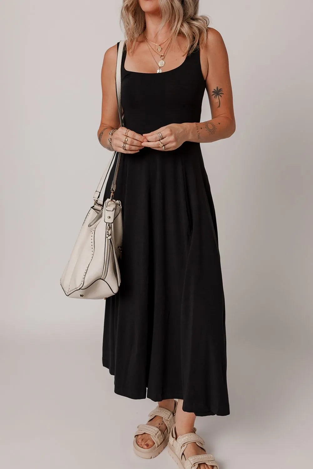 Woman wearing a black sleeveless dress with a white handbag on a plain background