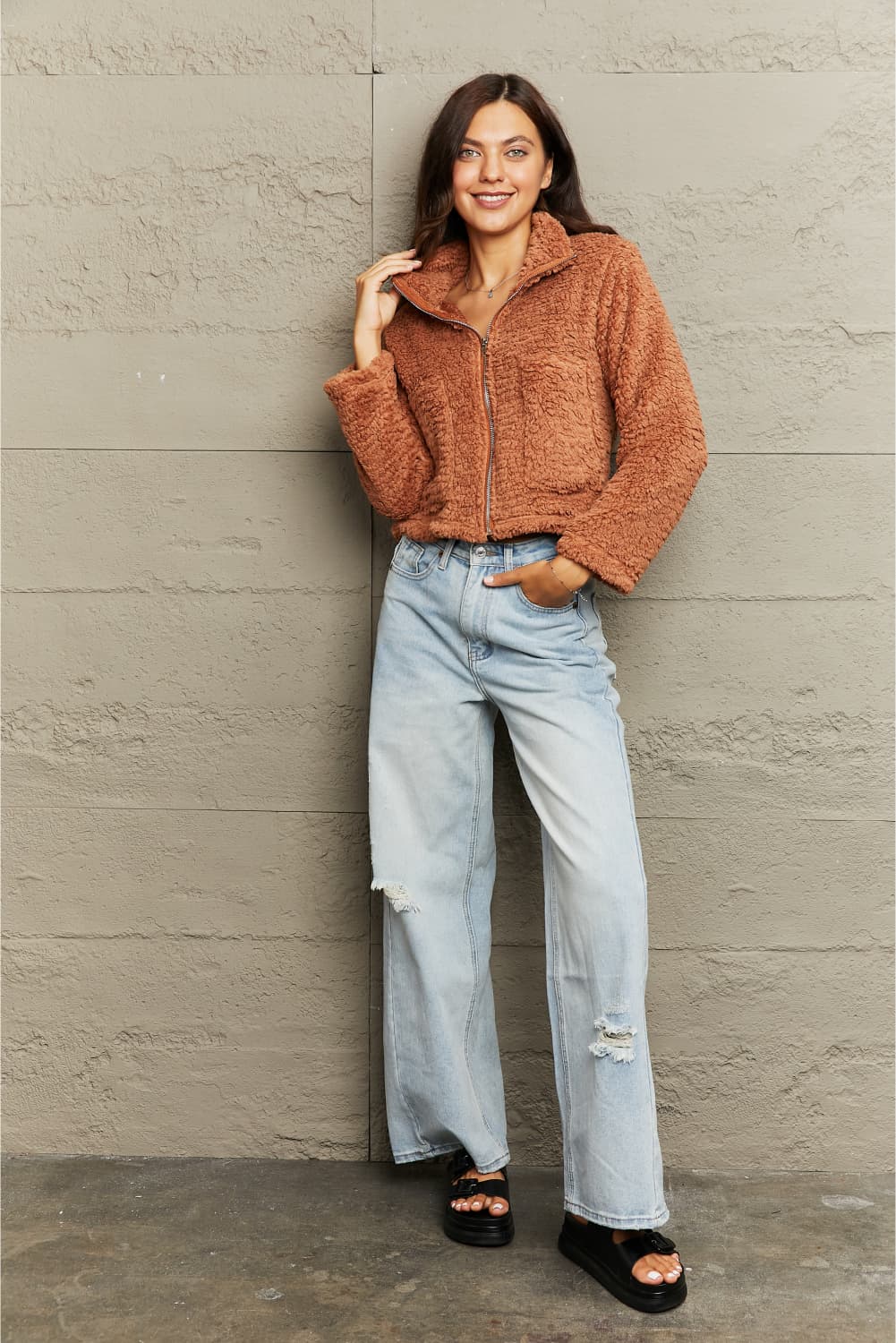 Woman wearing a brown knitted jacket and light blue jeans against a beige wall.