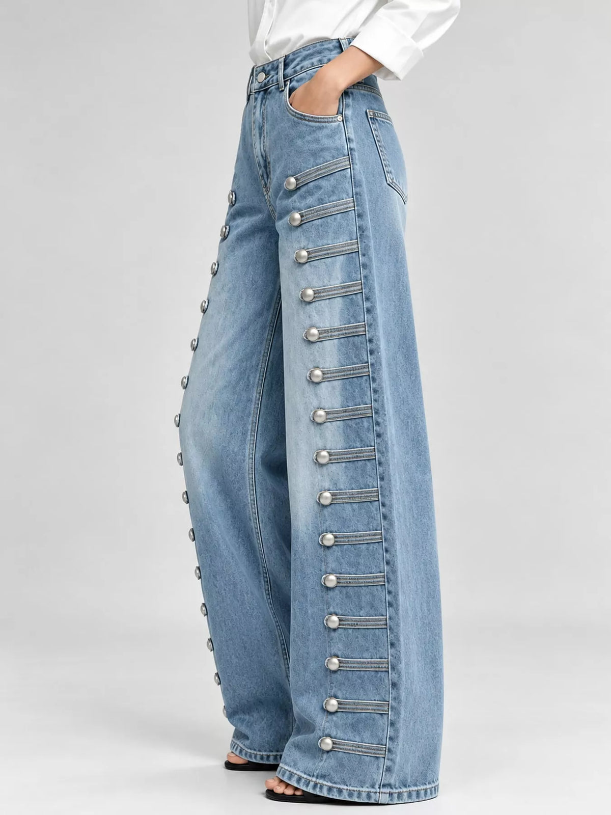 Light blue wide-leg denim jeans with decorative silver button and strap detailing down the outer side of each leg, shown on a model against a light gray studio background.