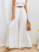 Person wearing a white outfit with wide-leg pants and a woven bag, standing against a plain background.