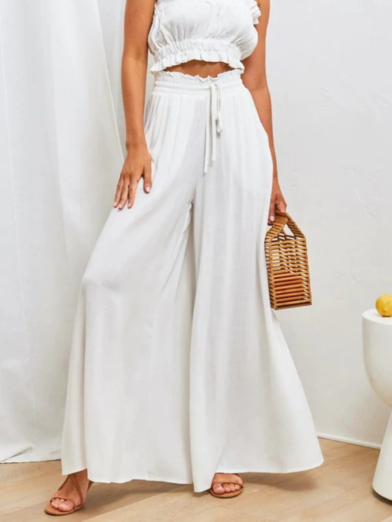 Person wearing a white outfit with wide-leg pants and a woven bag, standing against a plain background.
